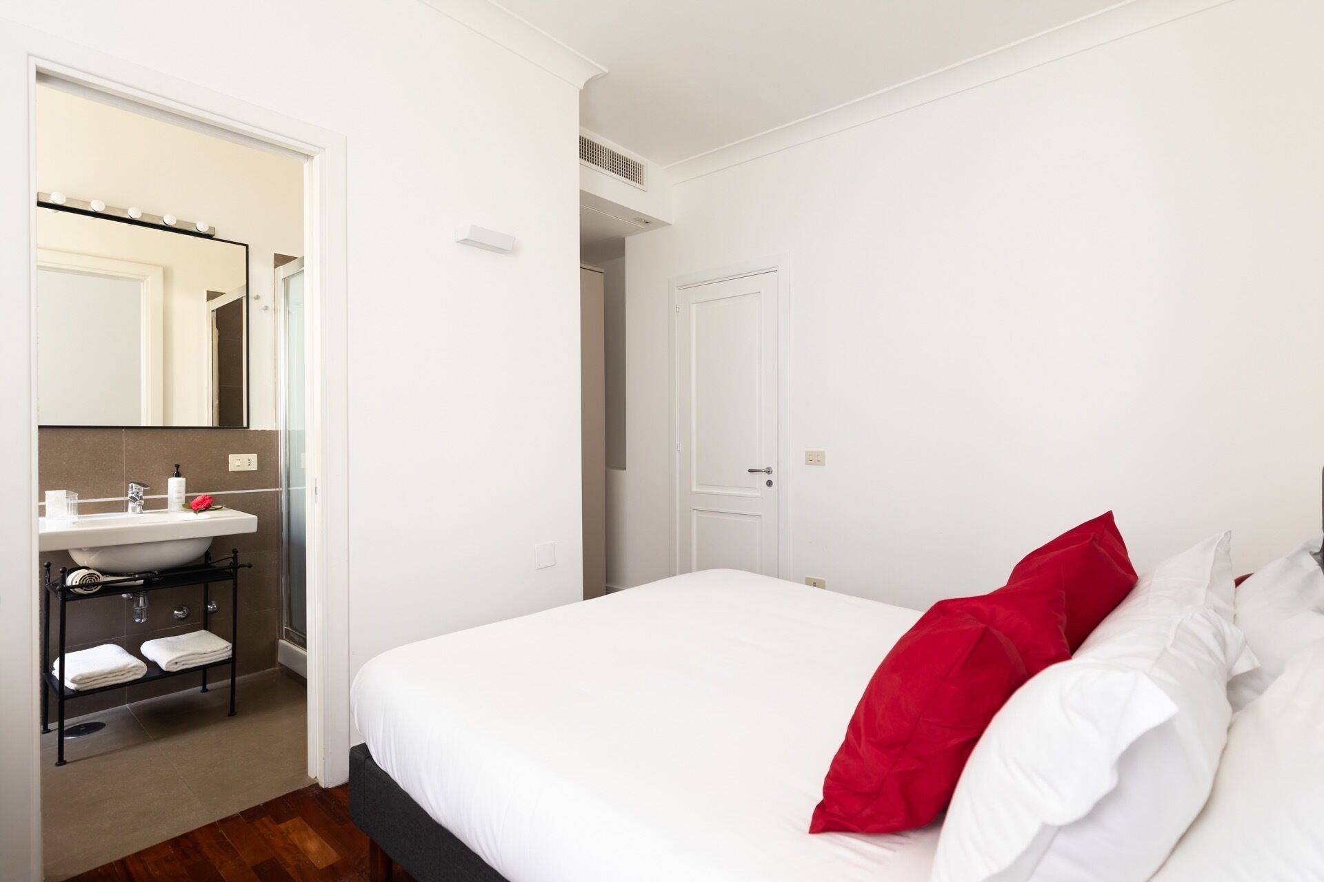 Una camera da letto con un letto, una parete bianca, una porta e un bagno con lavandino e specchio.