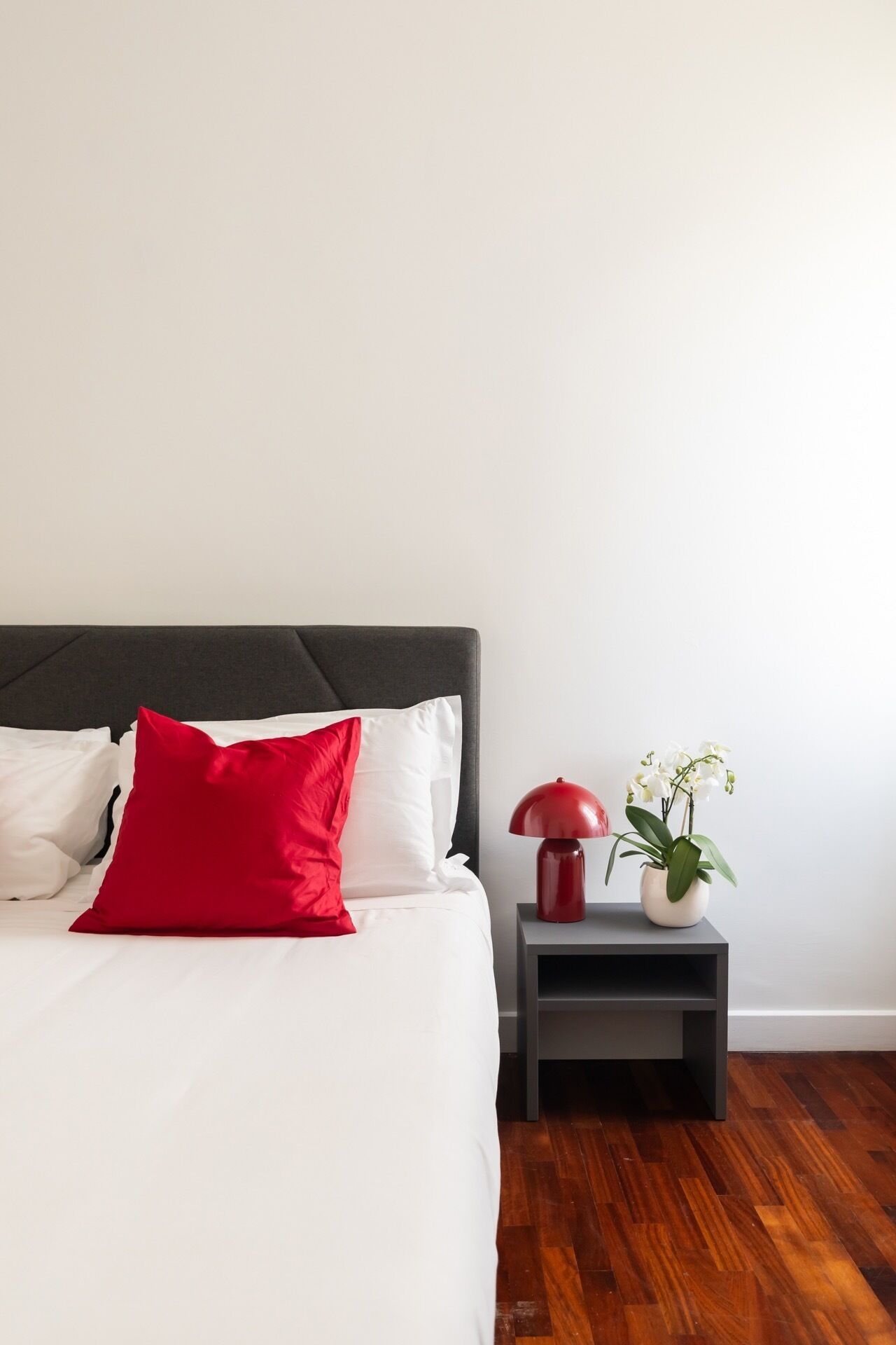 Un lit bien fait, agrémenté d’oreillers blancs et rouges, avec une tête de lit sombre et une petite table de chevet sur laquelle se trouvent une lampe rouge et une plante, une orchidée blanche.