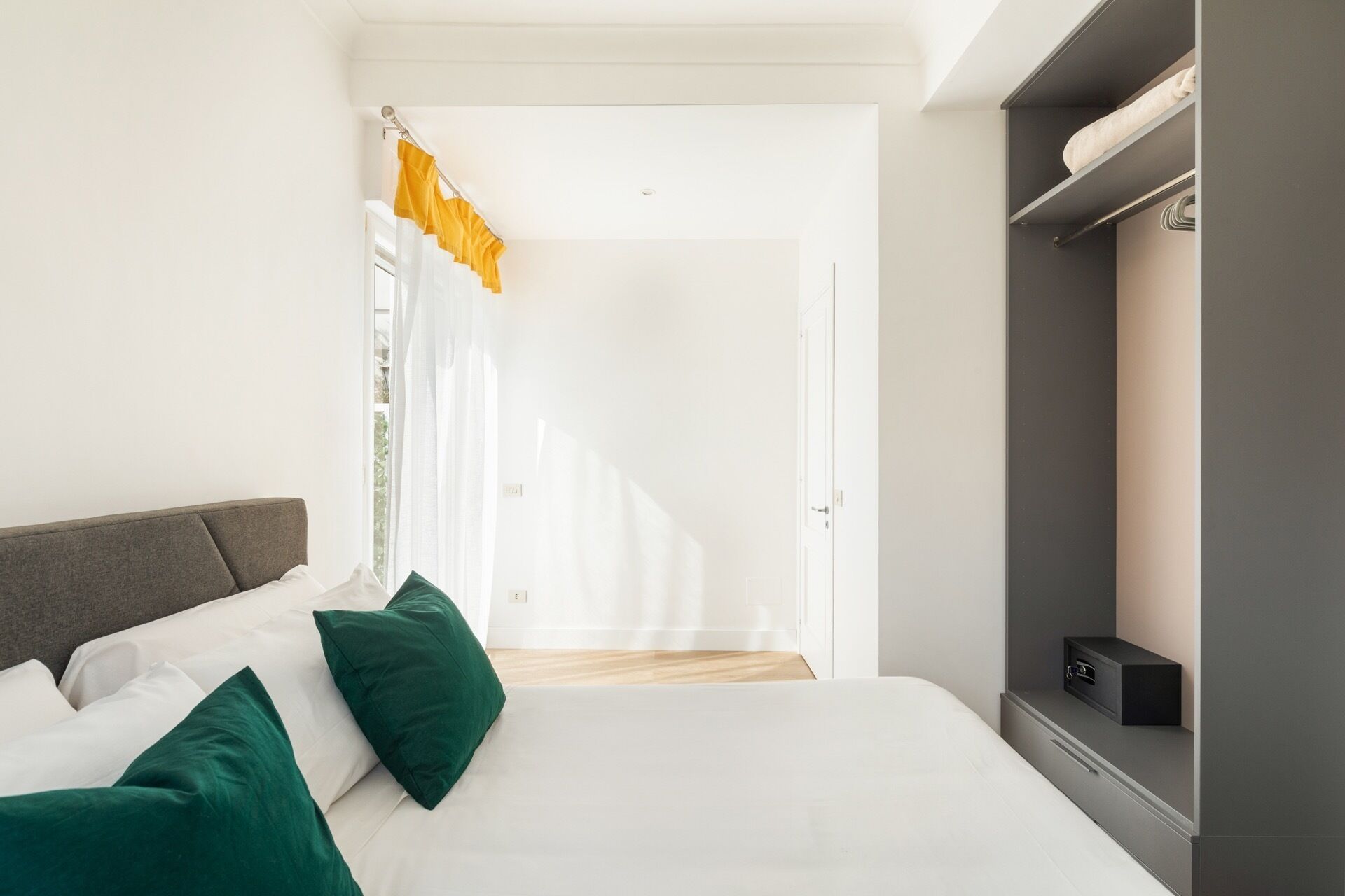 Un dormitorio moderno con una cama, almohadas verdes, un cabecero gris, un armario y una ventana con cortinas.