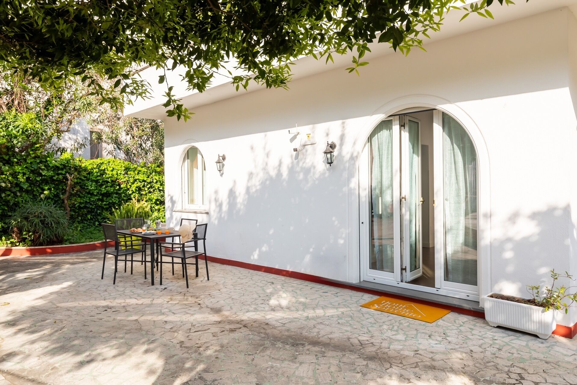 A white building with a covered patio, a table with chairs, and a potted plant.