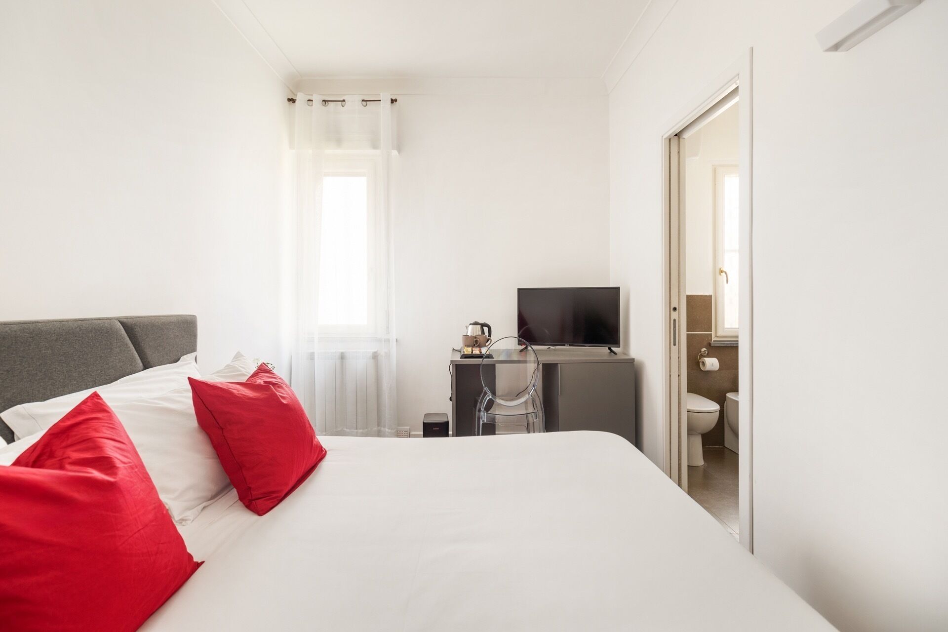 Habitación de hotel con cama, escritorio con televisor, ventana con cortinas translúcidas y vista al baño.