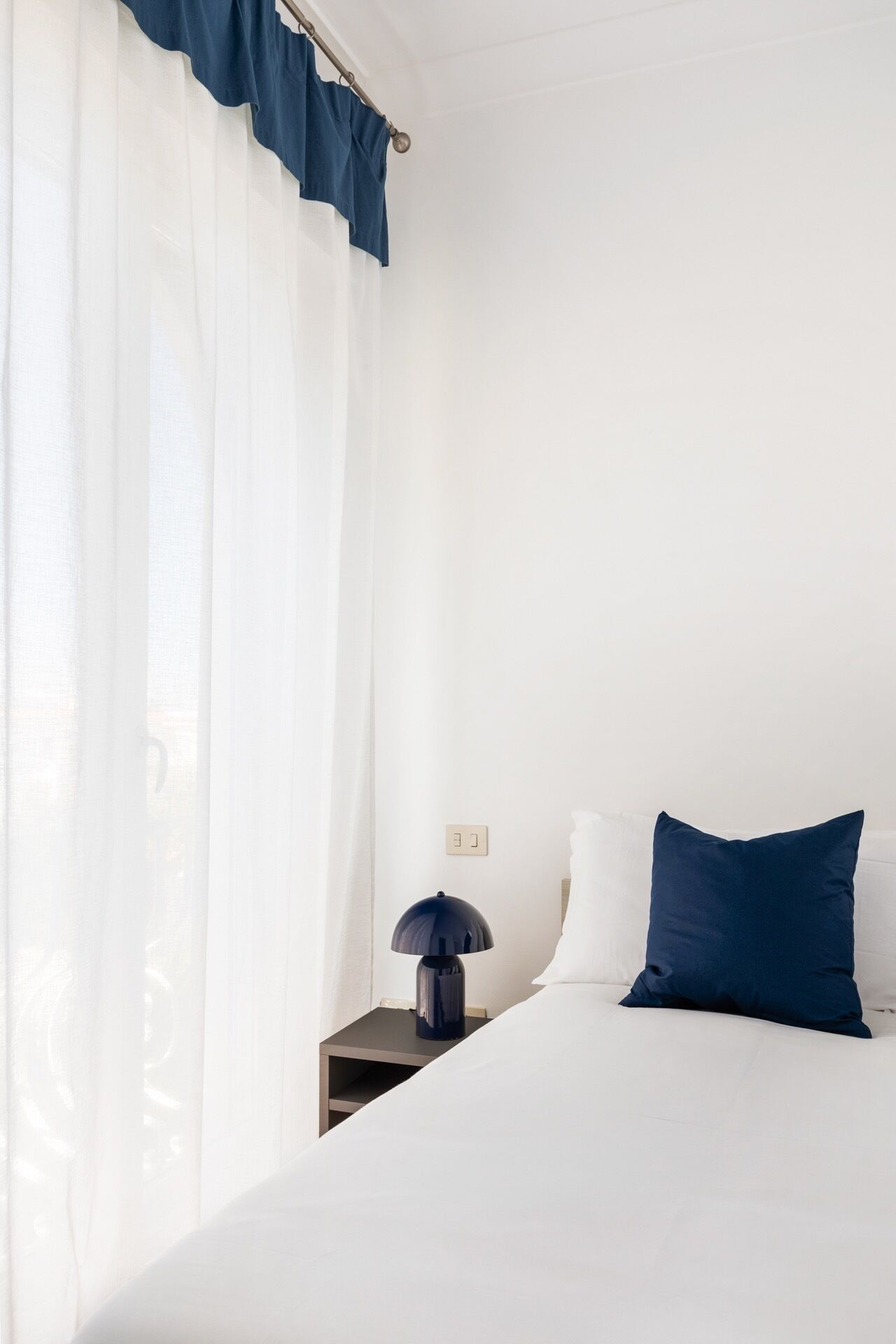 Ein Hotelzimmer mit Bett, Nachttisch, Lampe und transparenten Vorhängen.