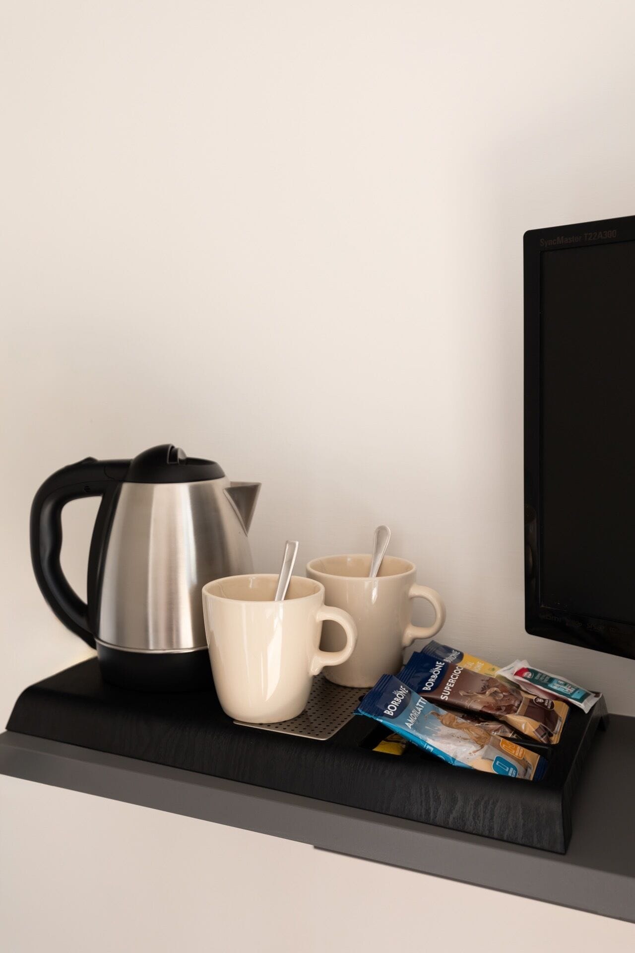 Een zwarte dienblad met een RVS-kettle, twee witte bekers met lepels, en een pak chocoladerepen op een plank.