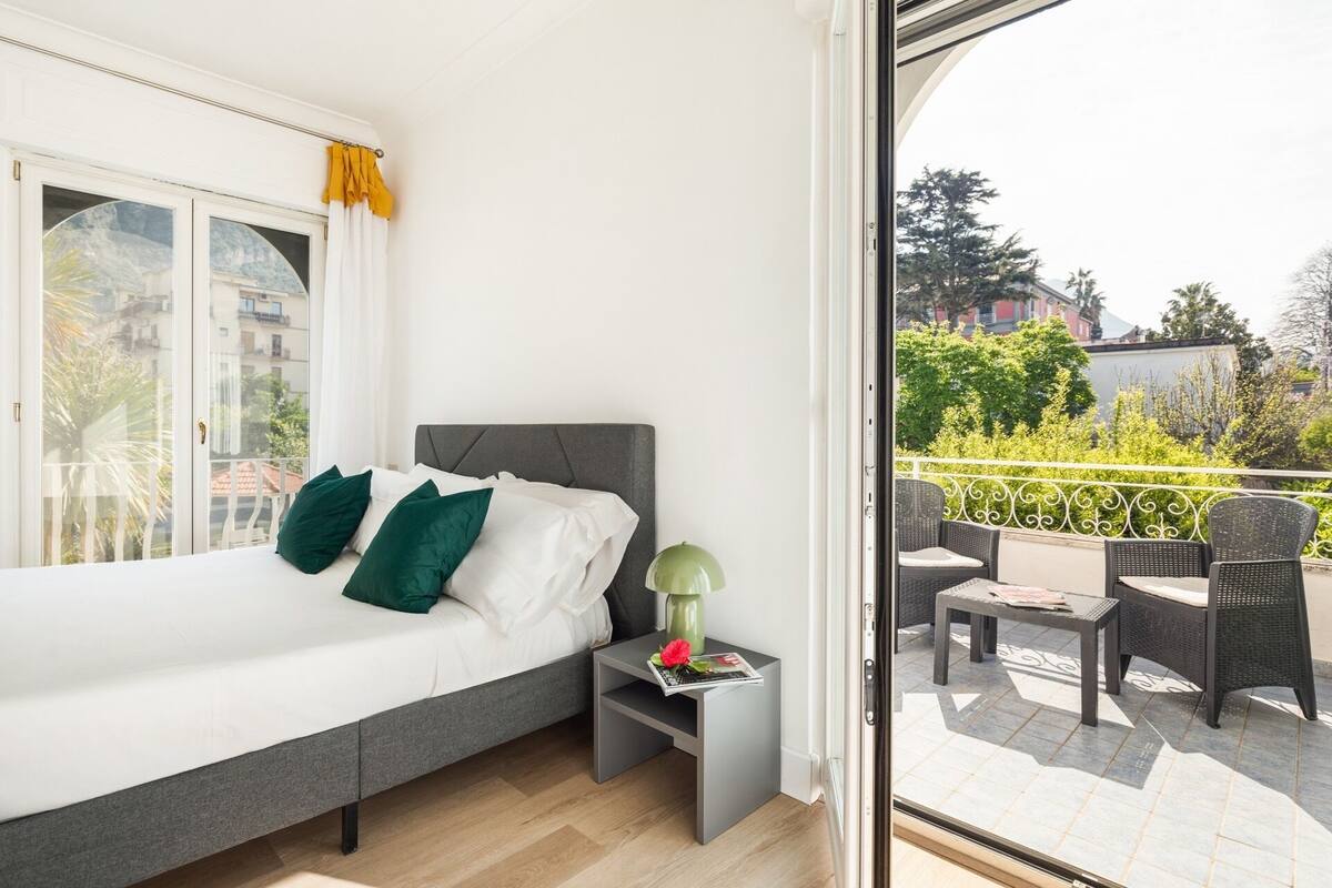 A bedroom with a bed, nightstand, and a view of a balcony with outdoor furniture.