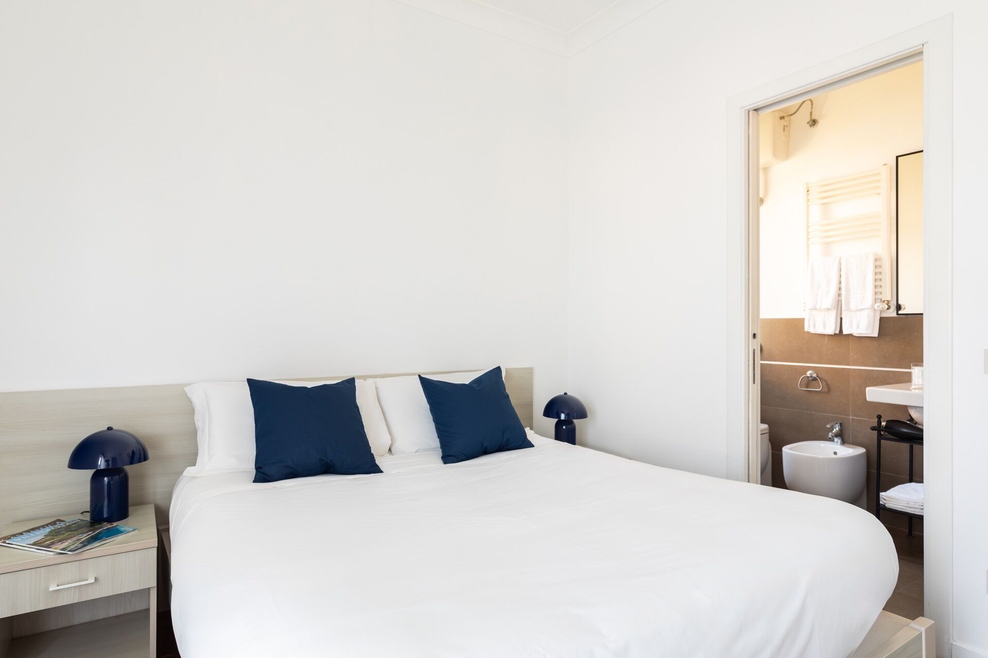 Una habitación de hotel con una cama grande, dos lámparas de noche, una mesita de noche con una revista, y un baño visible a través de una puerta abierta.