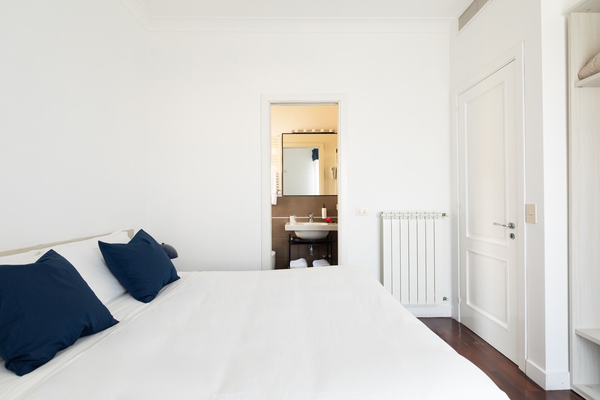 A bedroom with a large bed, two pillows, a nightstand, a mirror, and a bathroom visible through an open door.