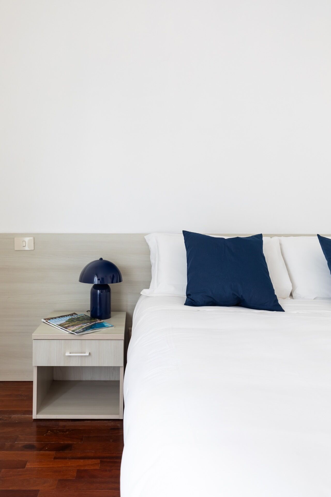 A neatly made bed with white and blue bedding, a bedside table with a lamp, and a magazine on the table.