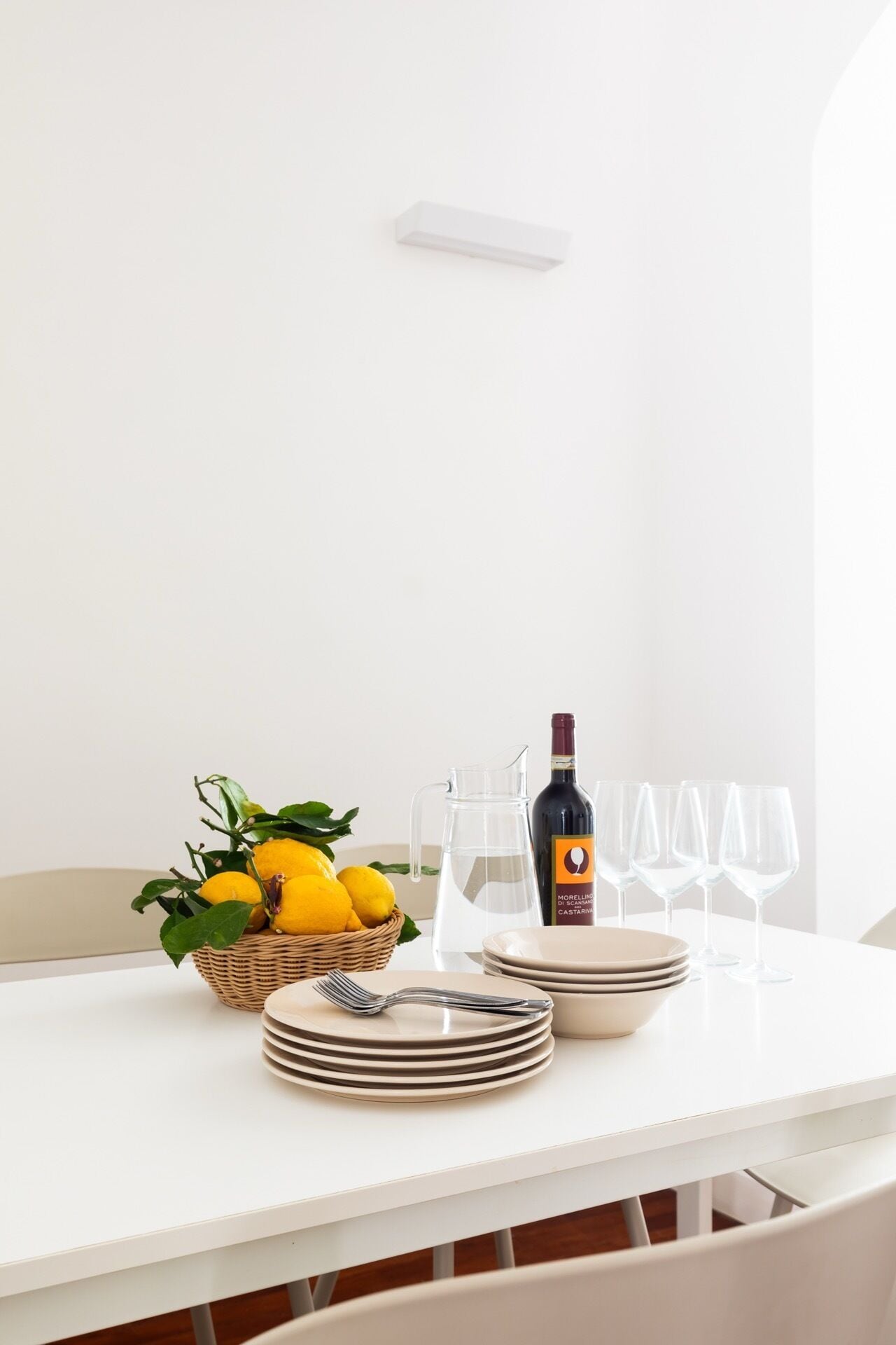 A white dining table with a basket of lemons, a wine bottle, and stacked plates and glasses.