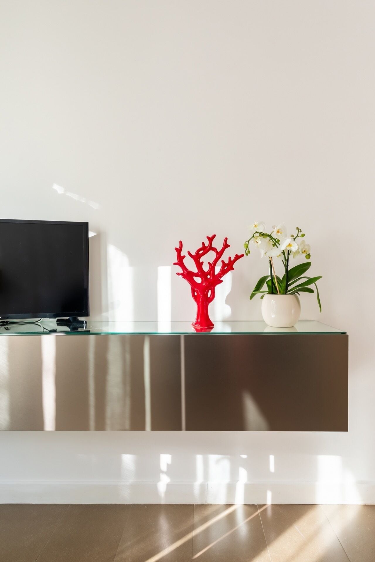 Un salon moderne avec un téléviseur à écran plat posé sur une étagère en verre, un vase rouge aux motifs abstraits et une plante d’orchidée blanche.