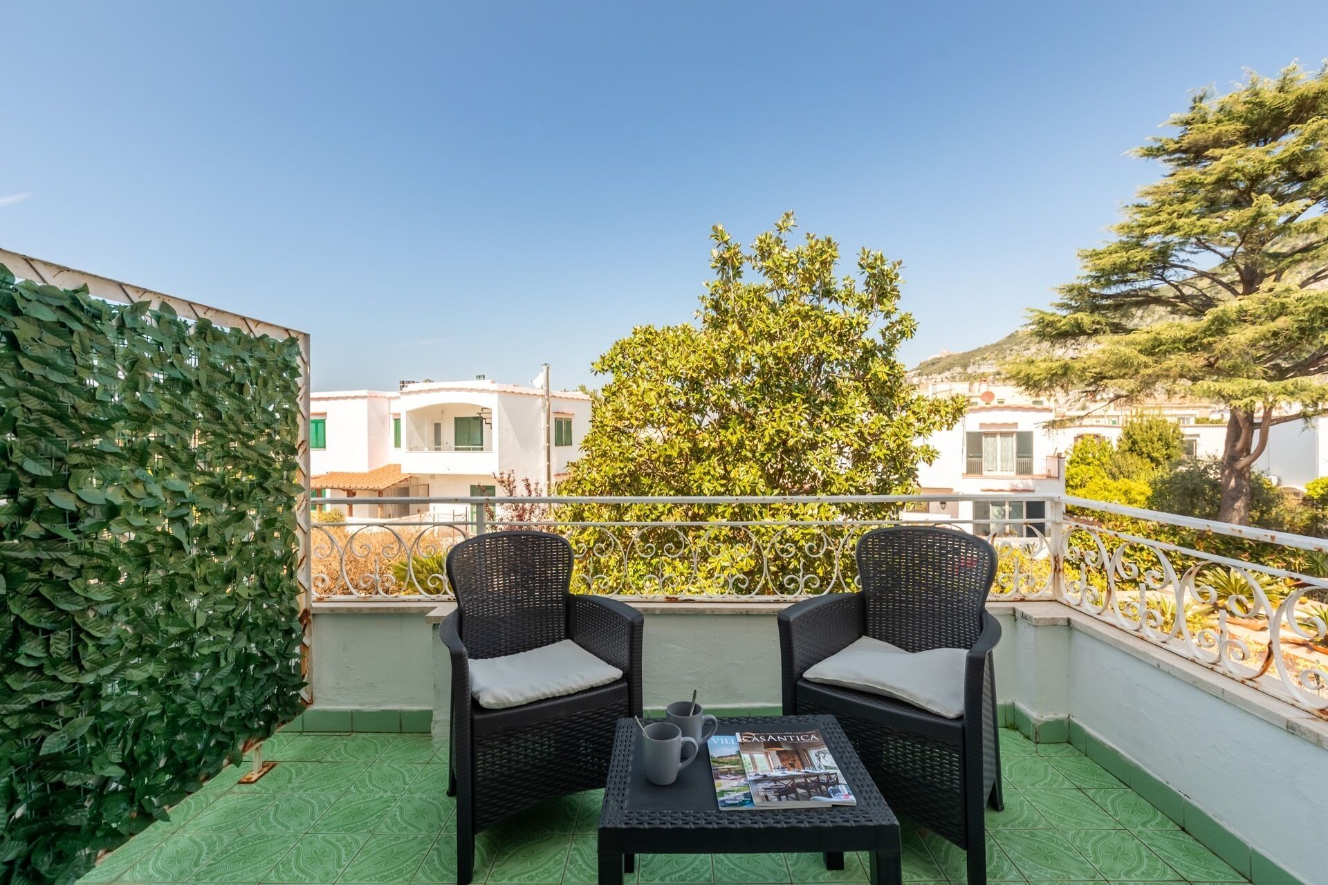 A balcony with two wicker chairs, a small table, and a magazine. The balcony has a green floor and a white railing.