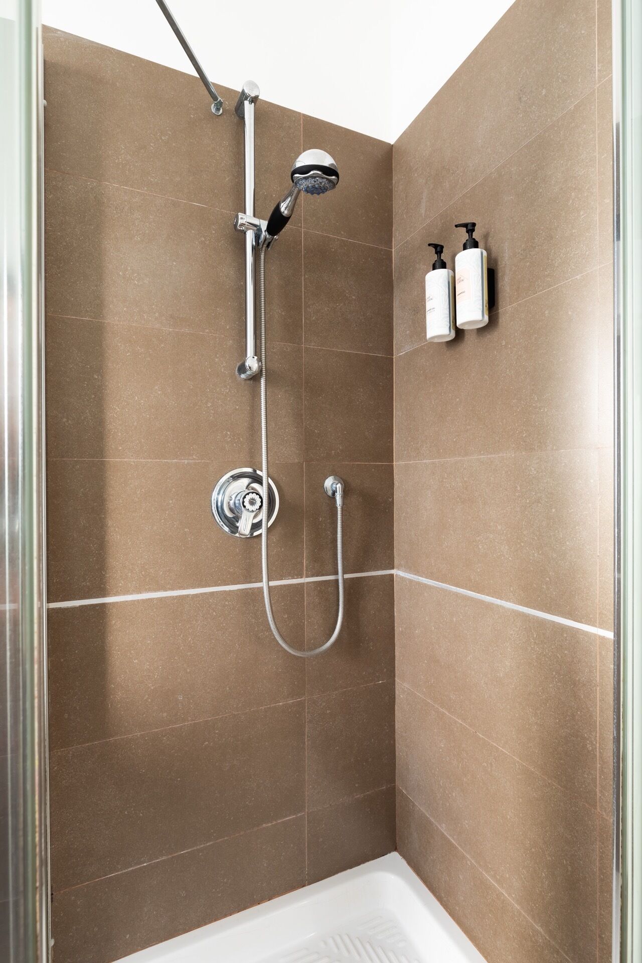 A shower with a handheld showerhead, a wall-mounted showerhead, and a handheld showerhead with a hose. There are two bottles of shampoo and conditioner on the wall.