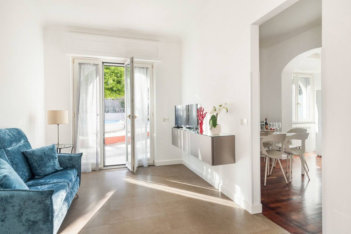 A modern living room with a blue sofa, a TV on a wall-mounted shelf, and a dining area with white chairs and a table.