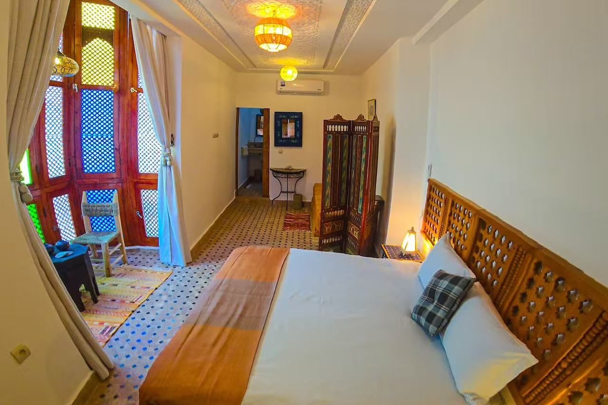 A room with a bed, a small table, a chair, a decorative rug, and a window with curtains.