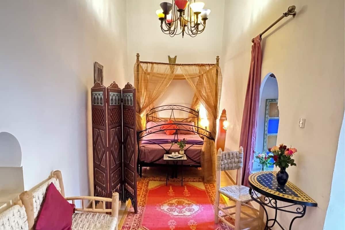 A bedroom with a bed, a sitting area with a sofa, a small table with a vase, and a decorative wall.