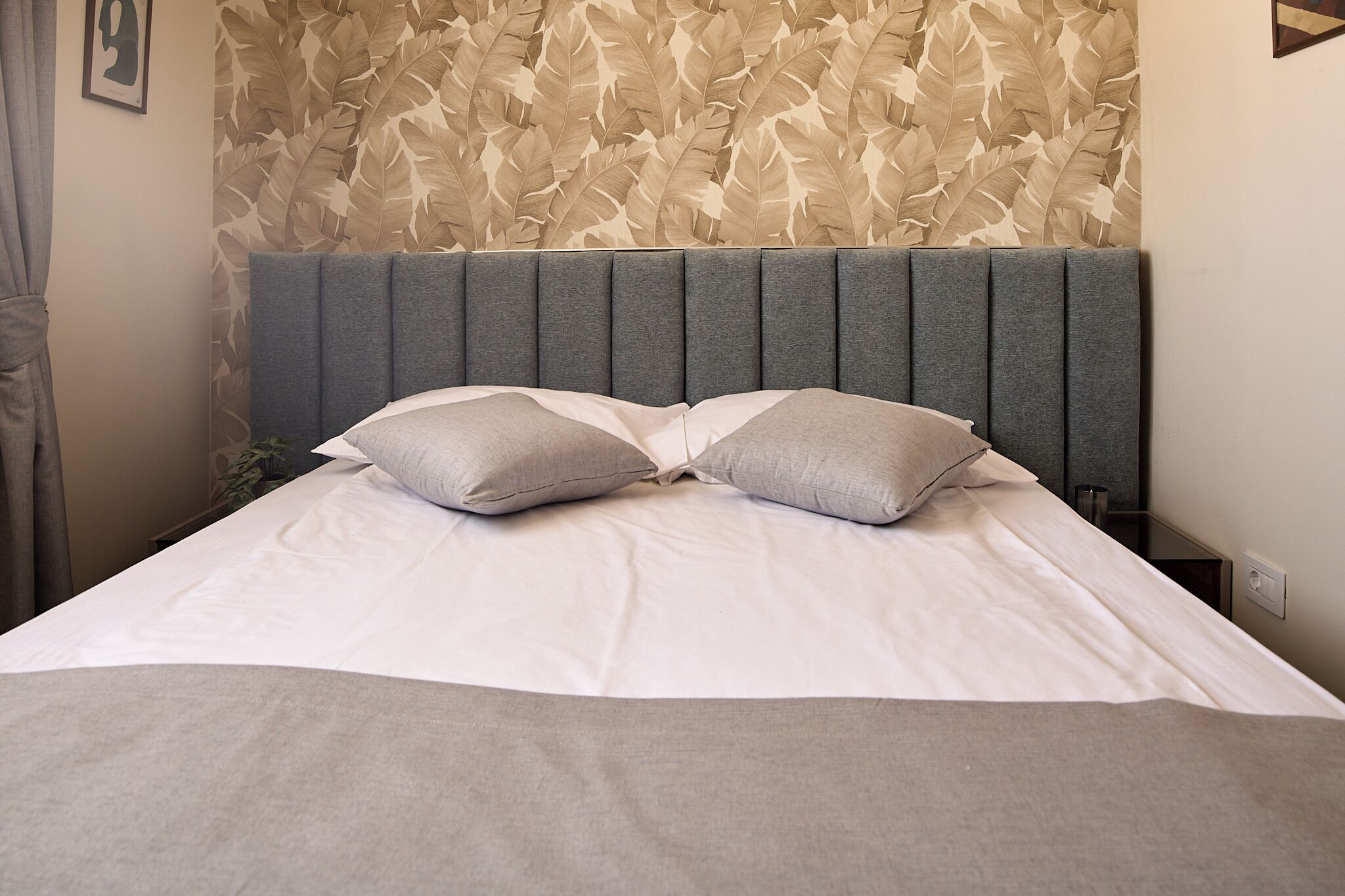 Cama bem arrumada com cabeceira cinzenta e dois travesseiros, parede com padrão de folhas e um quadro pendurado na parede.