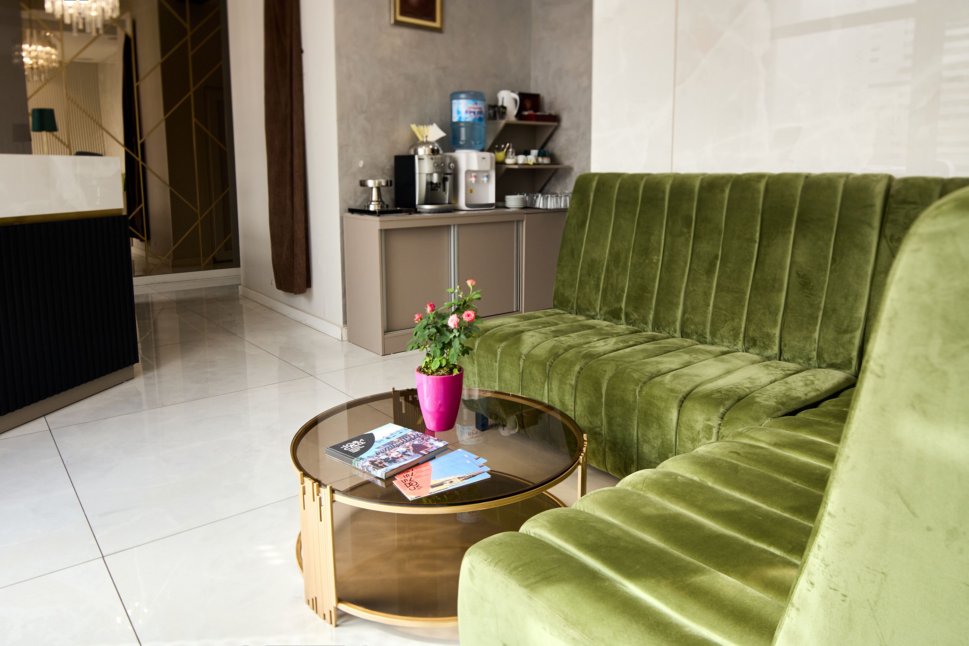 A modern living room with a green sofa, a glass coffee table with magazines, and a kitchen area in the background.
