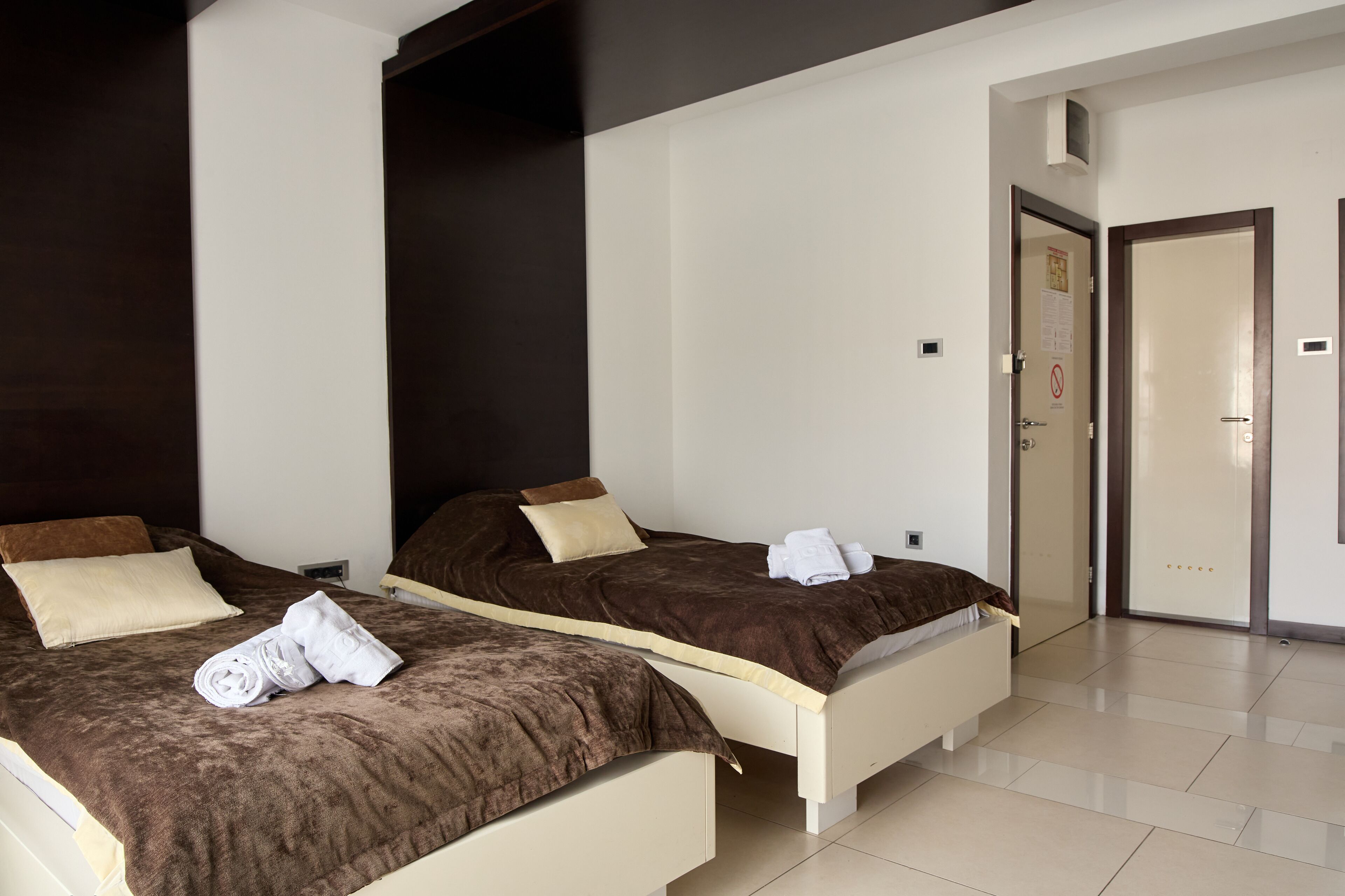 A hotel room with two beds, each with a brown headboard, white bedding, and a rolled towel on the bed.