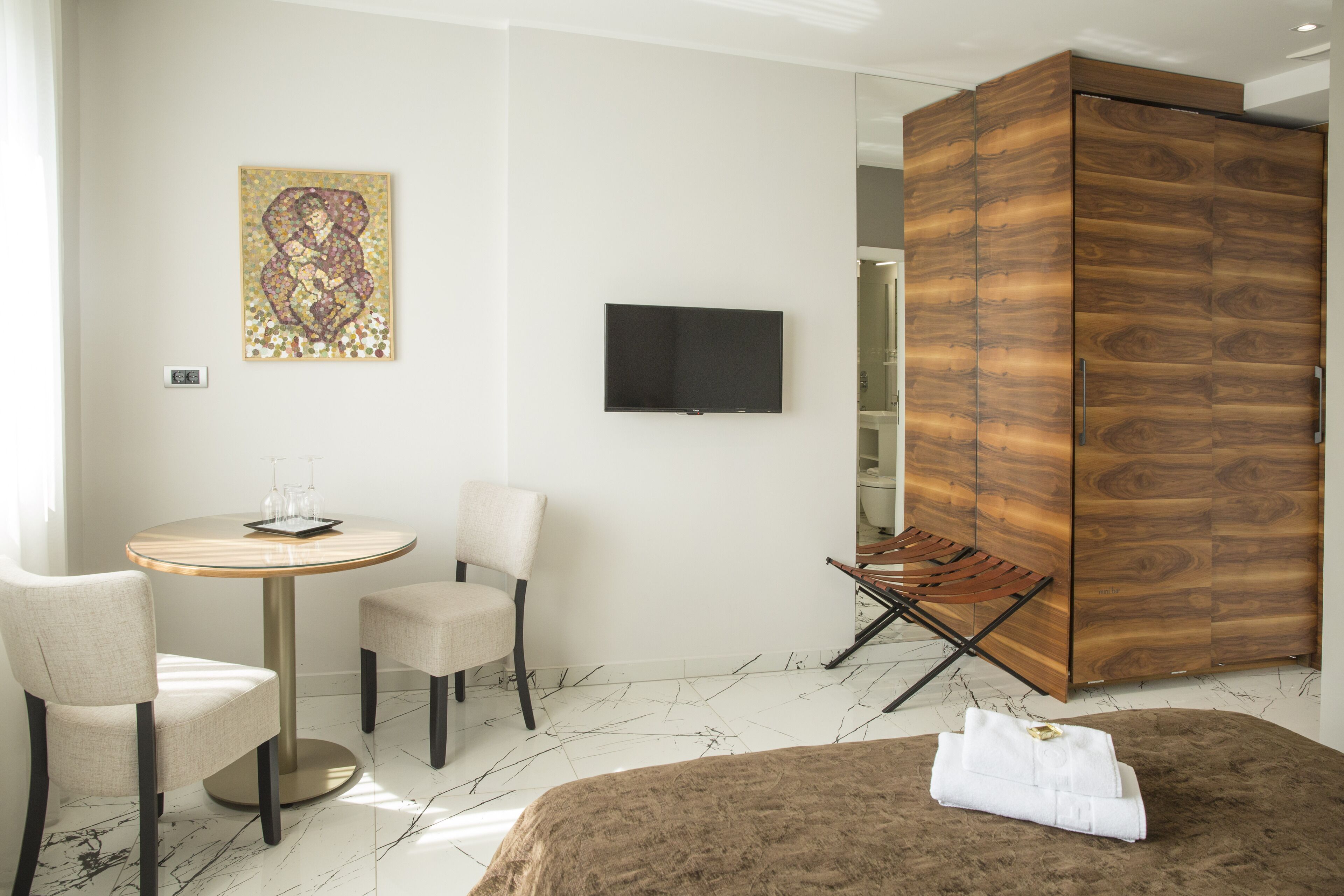 A hotel room with a bed, a round table with two chairs, a television, a wooden wardrobe, and a painting on the wall.
