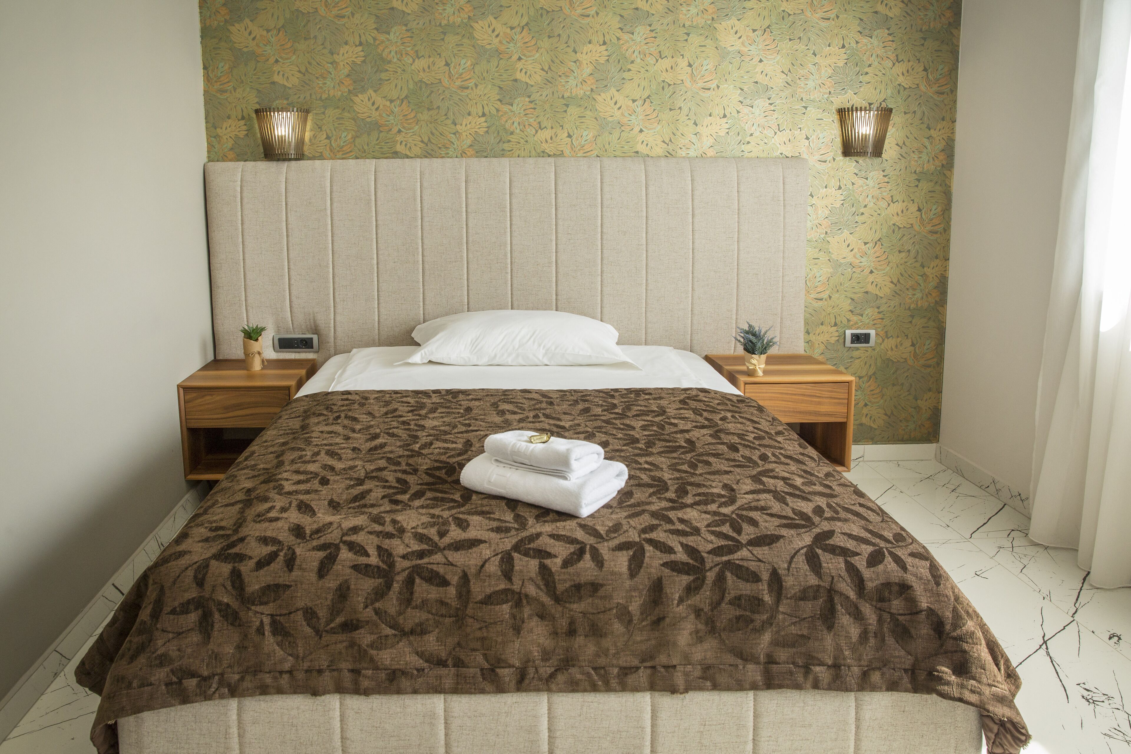 Un lit bien fait, avec une couvre-lit à motifs et une serviette pliée posée sur le côté. Il y a deux tables de chevet avec des lampes et un luminaire fixé au mur.