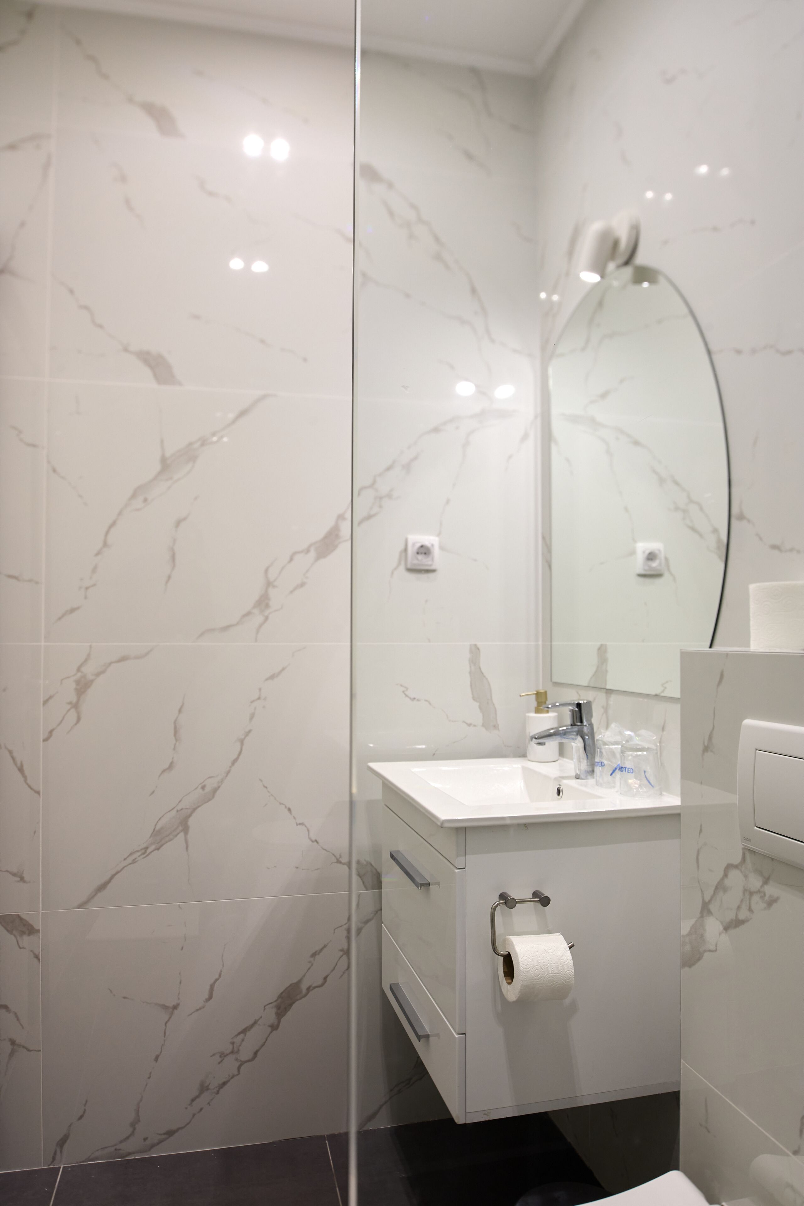 A modern bathroom with marble walls, a white vanity, a glass shower enclosure, and a toilet paper holder.