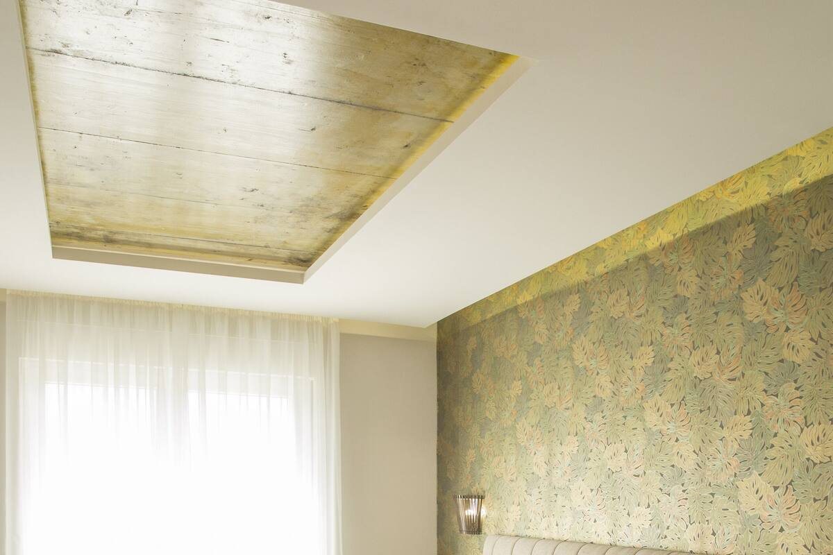 A hotel room with a bed, a bedside table, a chair, a window with curtains, and a patterned wall.