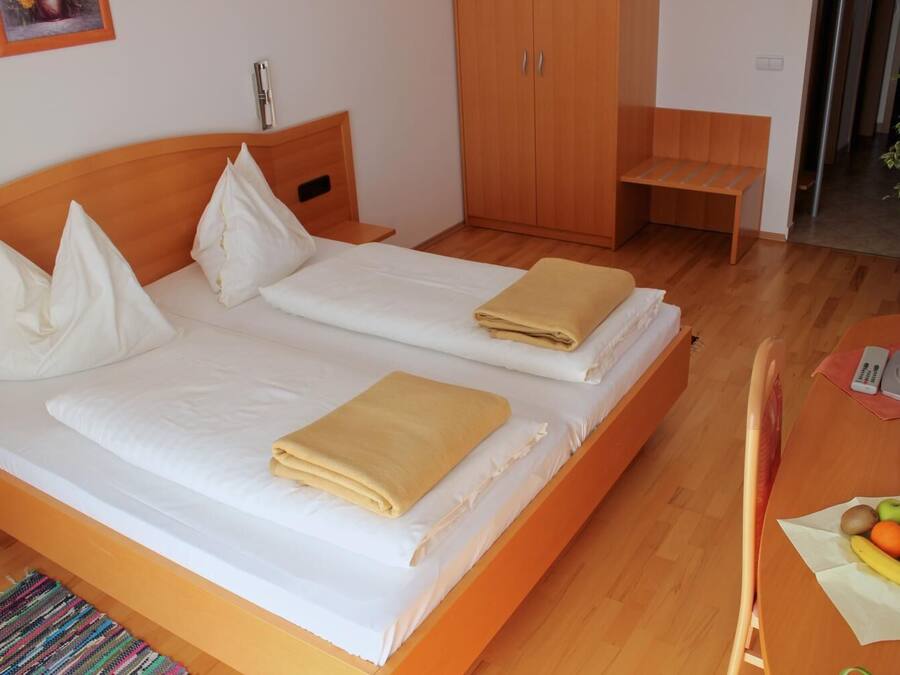 A double bed with white linens and two beige blankets, a wooden wardrobe, a bench, and a small table with fruits.