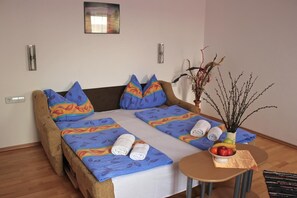 Una cama doble con ropa de cama colorida, una mesita redonda con fruta y un florero, y una lámpara fijada a la pared.