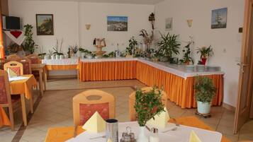 Un comedor con mesas dispuestas para una comida, rodeado de sillas. Se observan plantas en macetas y una mesa de buffet al fondo.