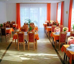 Un comedor con varias mesas y sillas dispuestas en filas, con cortinas naranjas y un ventanal grande al fondo.