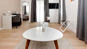Una habitación de hotel moderna con una mesa redonda blanca, un escritorio con teléfono y lámpara, una cama con cabecera oscura y un televisor en la pared.