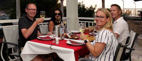 A group of people dining at an outdoor table with a red tablecloth, white chairs, and a view of the outdoors.