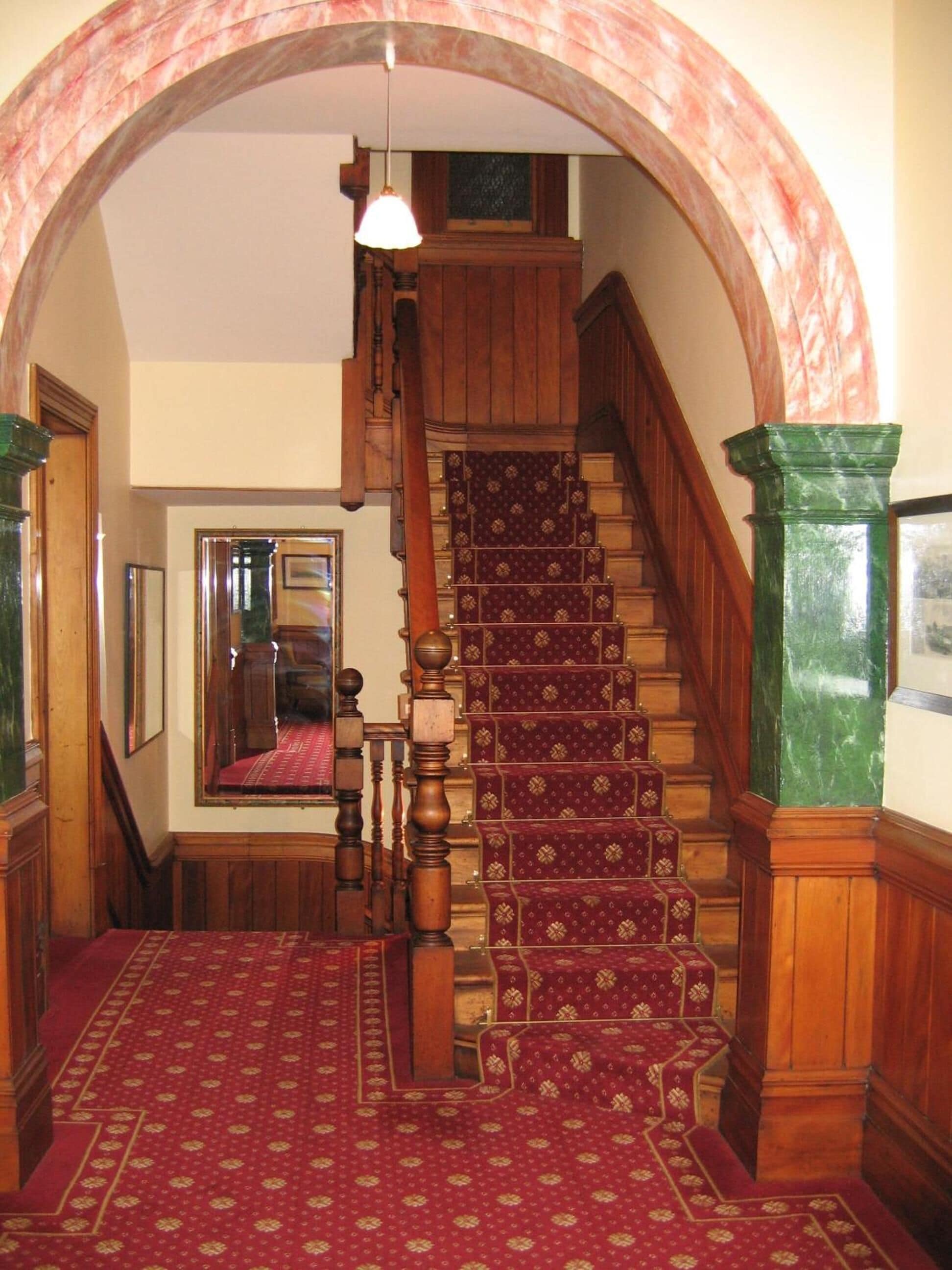 A grand staircase with a red carpet, wooden balusters, and a green marble column.