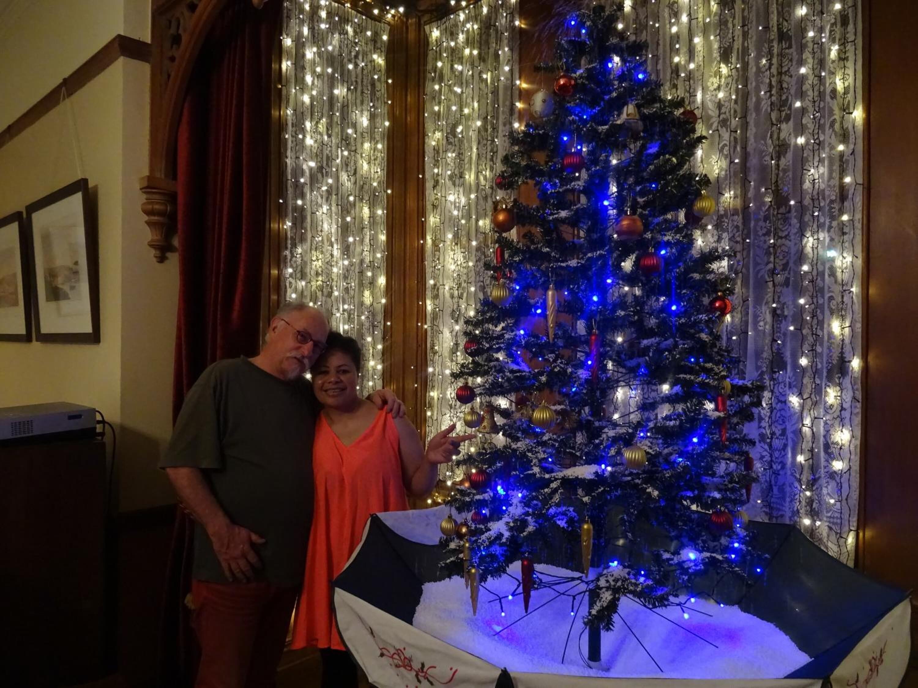 Een versierde kerstboom met lichtjes en decoraties, een persoon die er naast staat, en een gordijn met lichtslingers op de achtergrond.