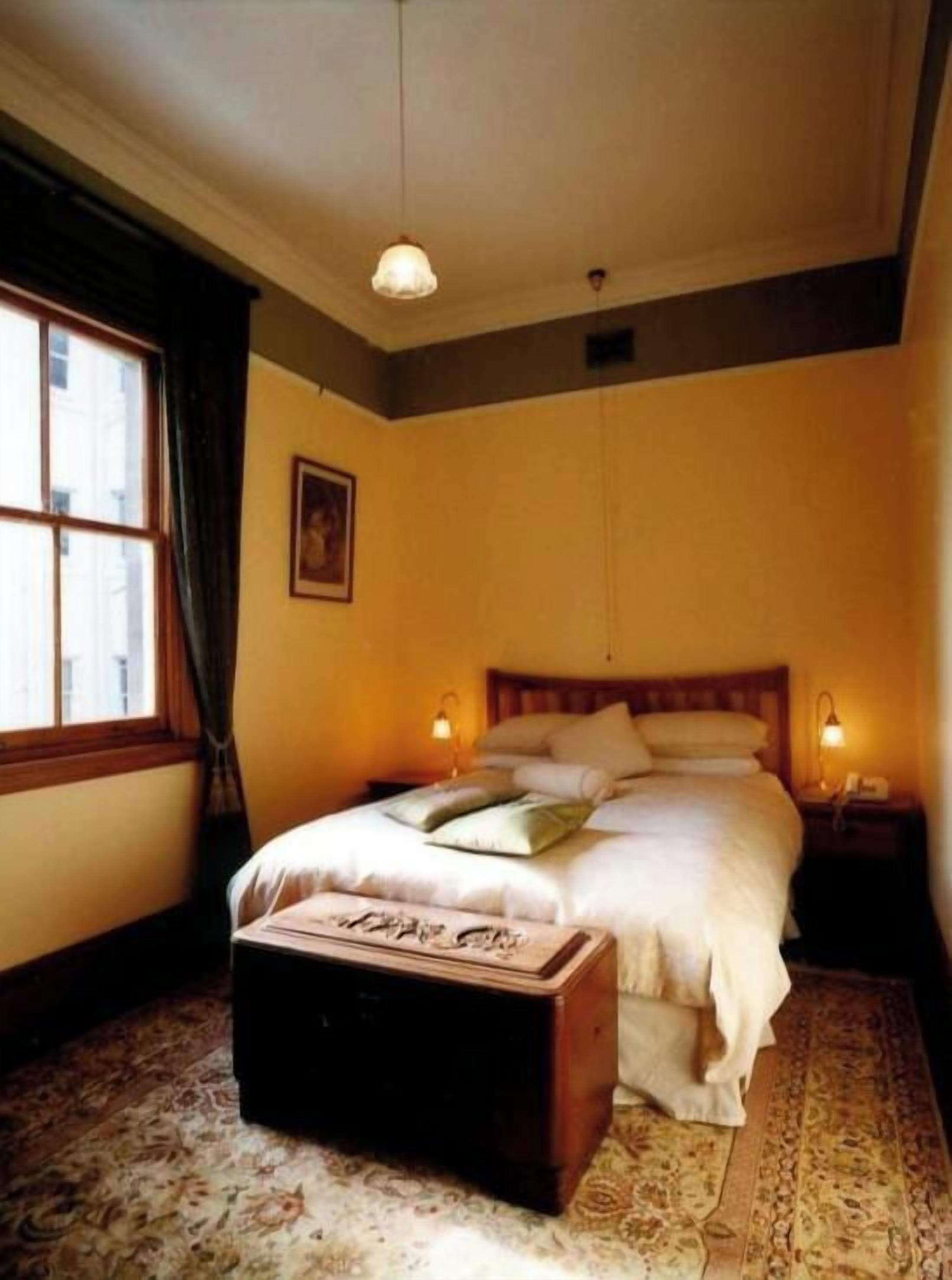 Un dormitorio con cama, lámparas de noche, mesita de noche, una ventana con cortinas y una alfombra estampada.