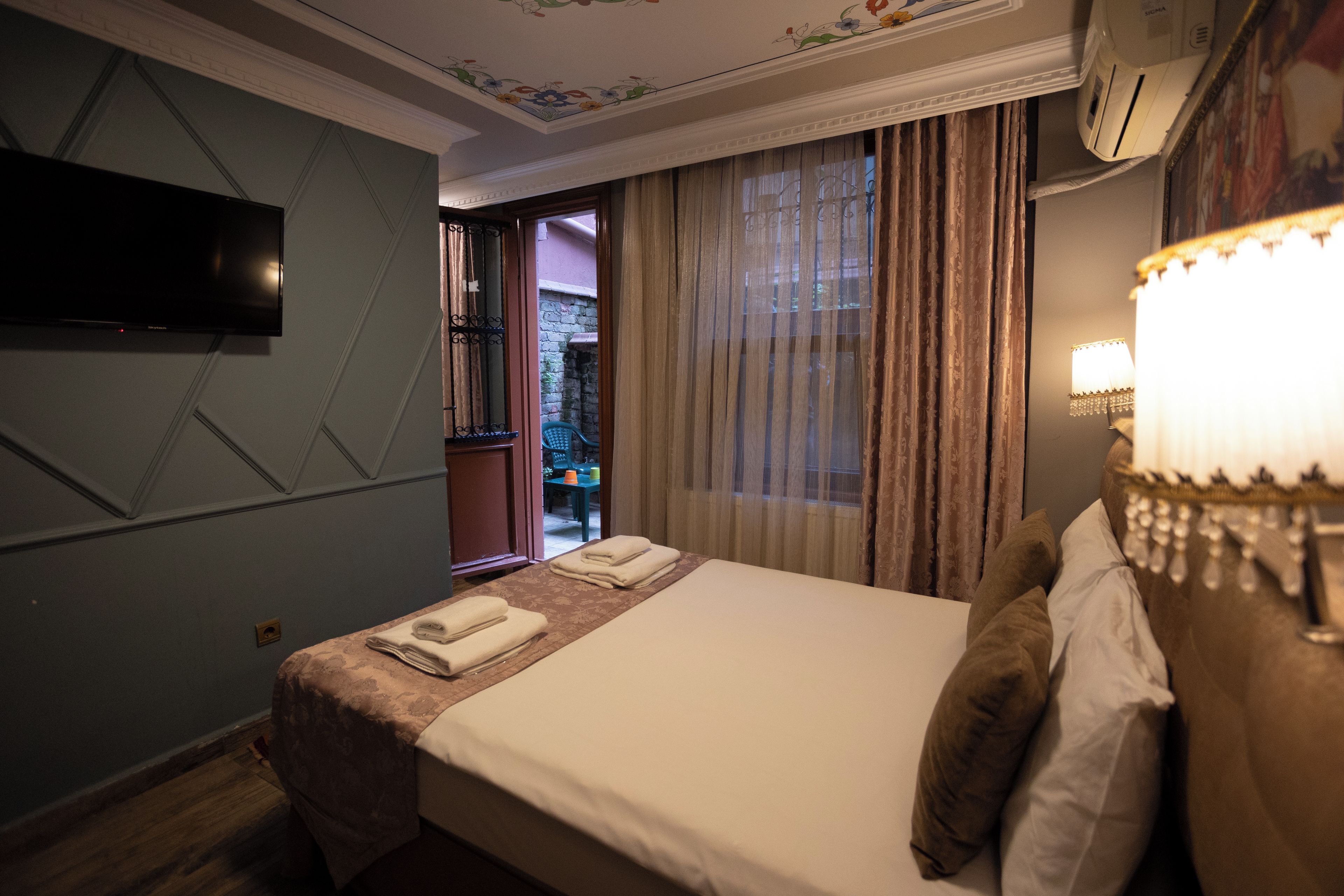 A hotel room with a bed, a flat-screen TV, a window with curtains, and a wall-mounted air conditioning unit.