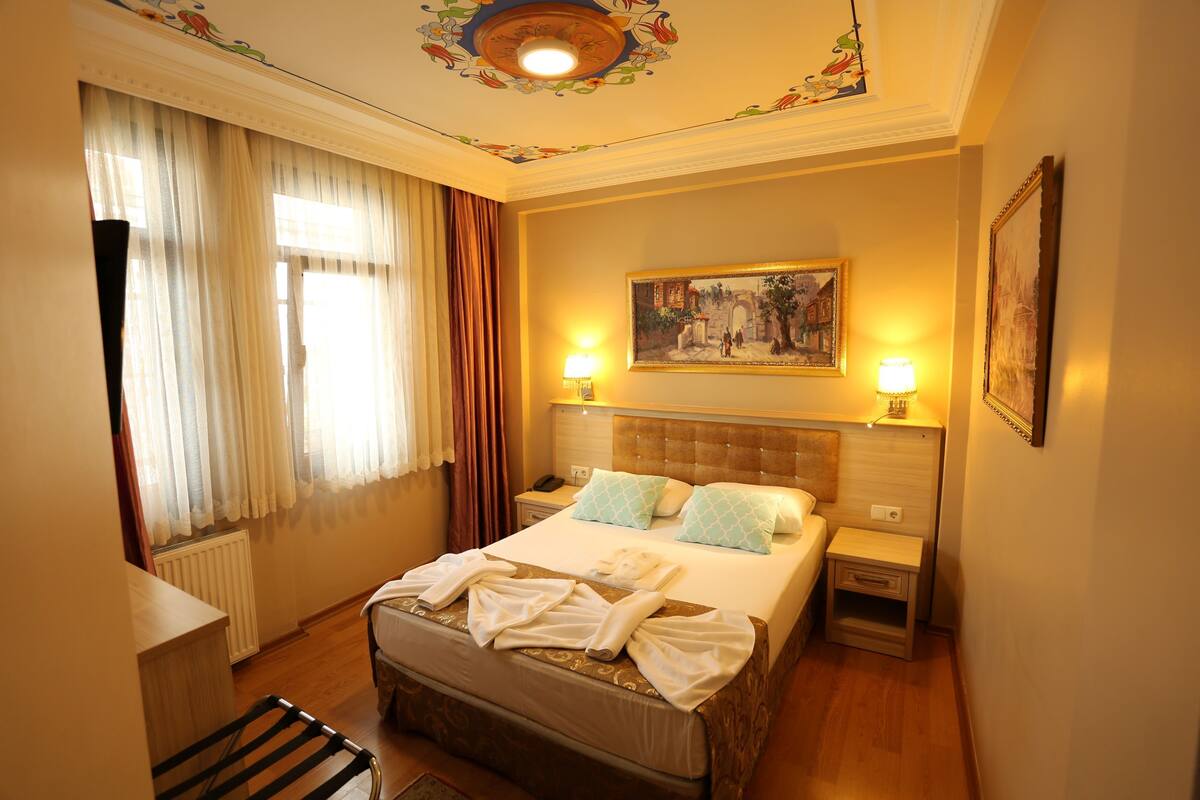 A hotel room with a large bed, two bedside tables with lamps, a painting on the wall, and a window with curtains.