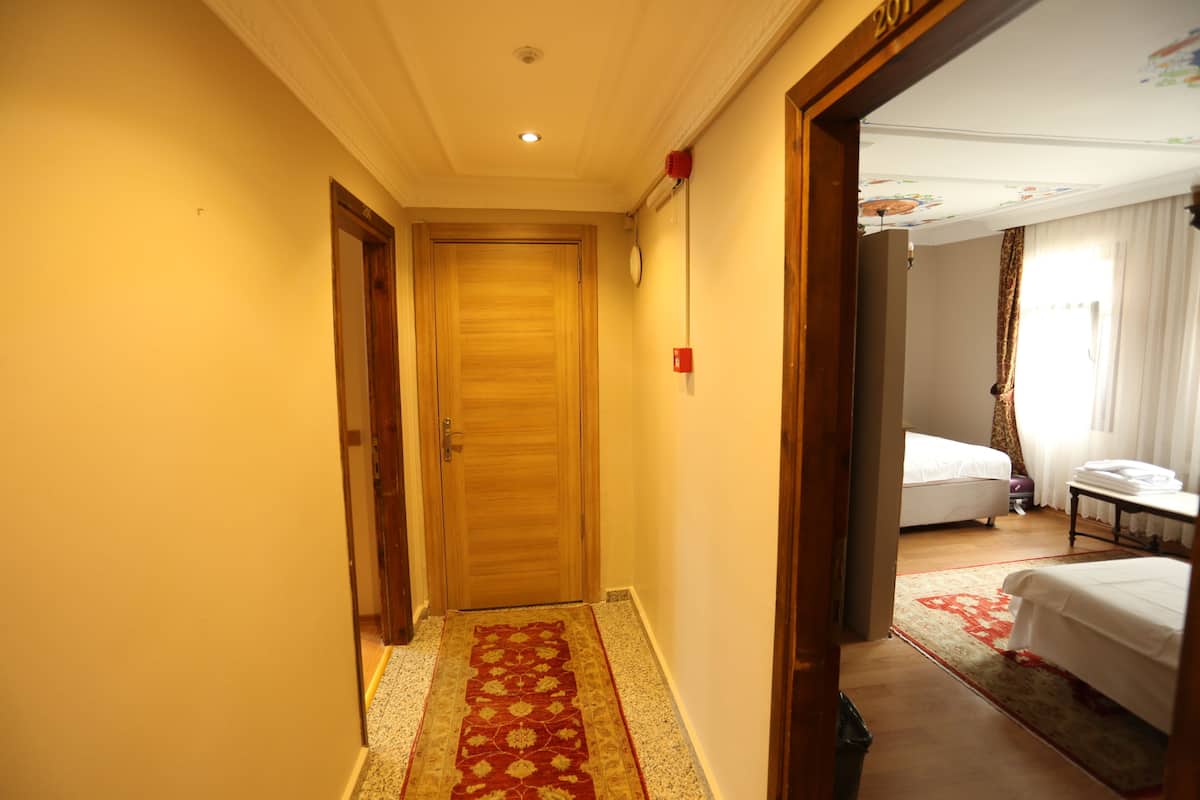 A hotel hallway with a wooden door, a red patterned carpet, and a bed with white bedding in the adjacent room.