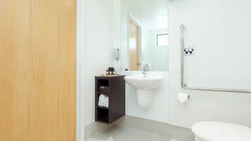 A modern bathroom with a wooden door, a dark cabinet, a white sink, a mirror, a handrail, and a toilet.