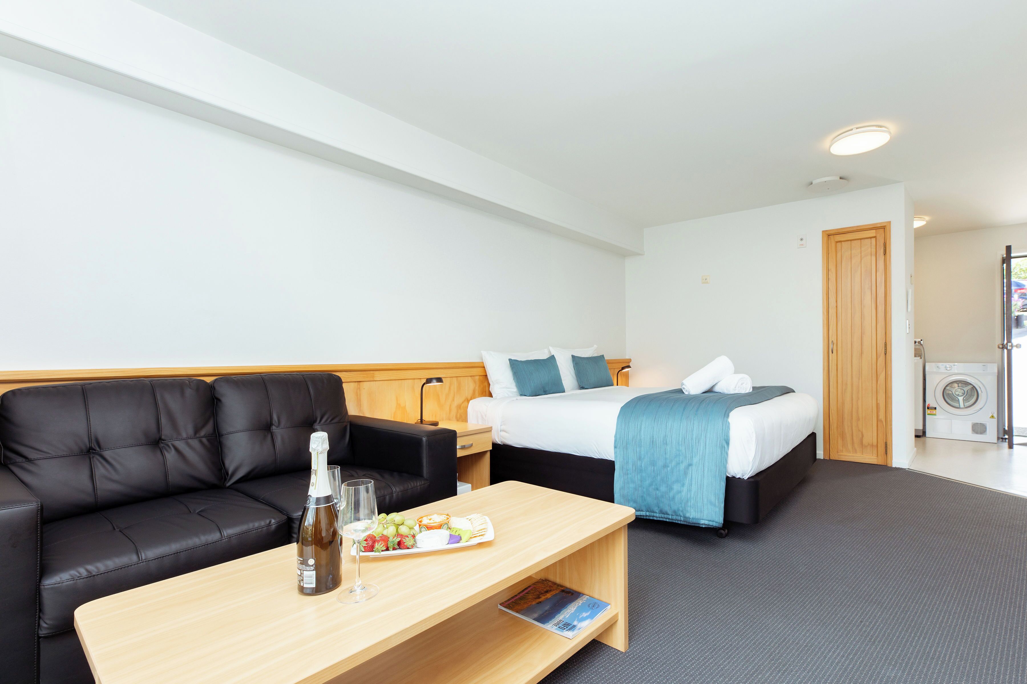 A hotel room with a black leather sofa, a wooden coffee table with a bottle and glass, a bed with a blue blanket, and a washing machine in the corner.