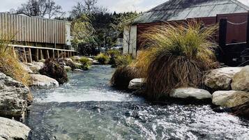 A clear stream flowing through a natural landscape with rocks and vegetation, a wooden structure, and a building with a metal roof.