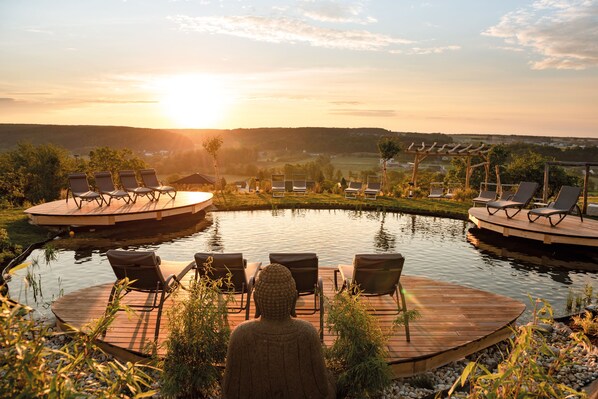 A serene outdoor pool area with lounge chairs and a Buddha statue, surrounded by a wooden deck and overlooking a scenic landscape during sunset. - Hotel & Spa Larimar (Stegersbach)