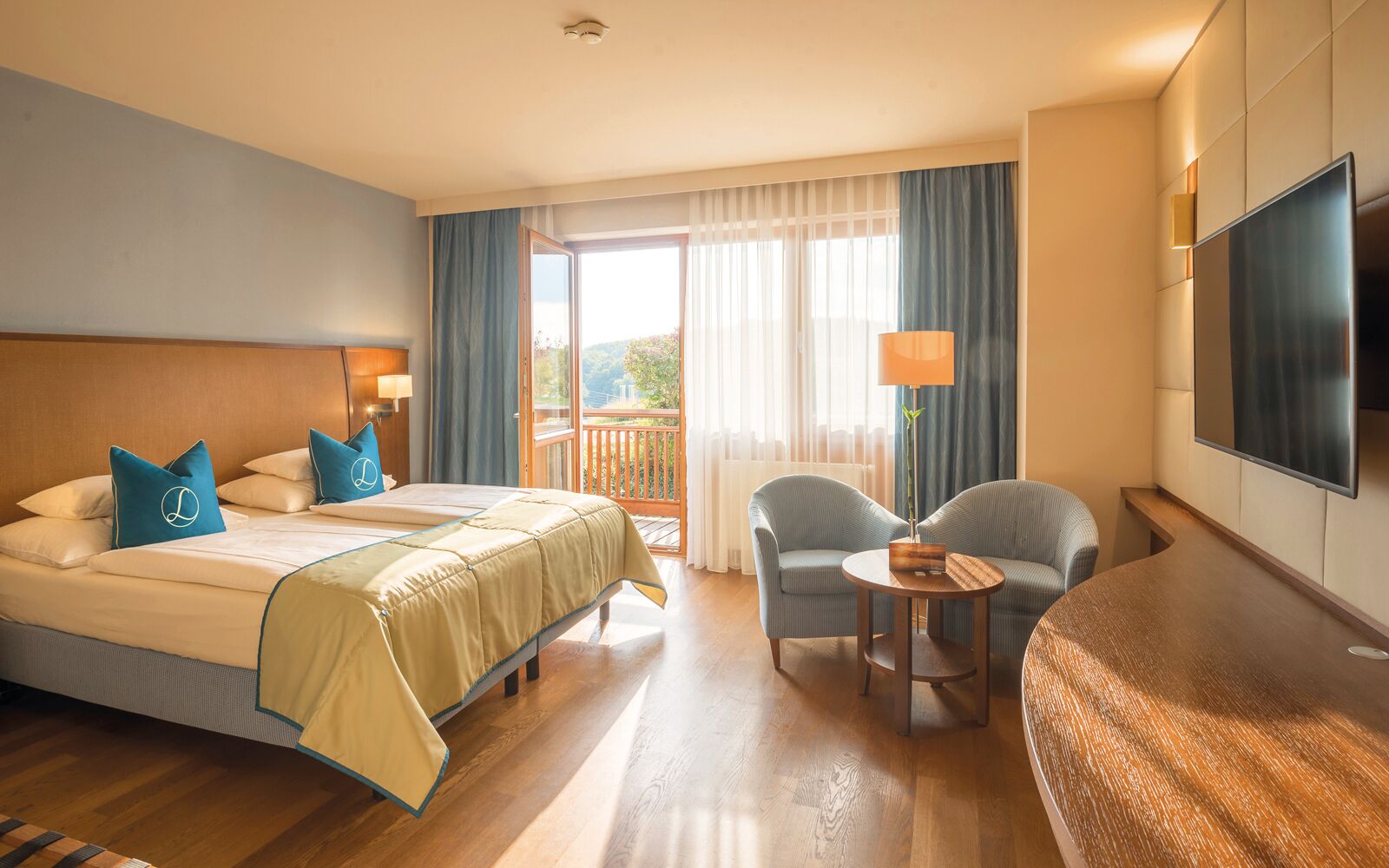 A hotel room with a bed, two armchairs, a small table, a TV, and a balcony with curtains.
