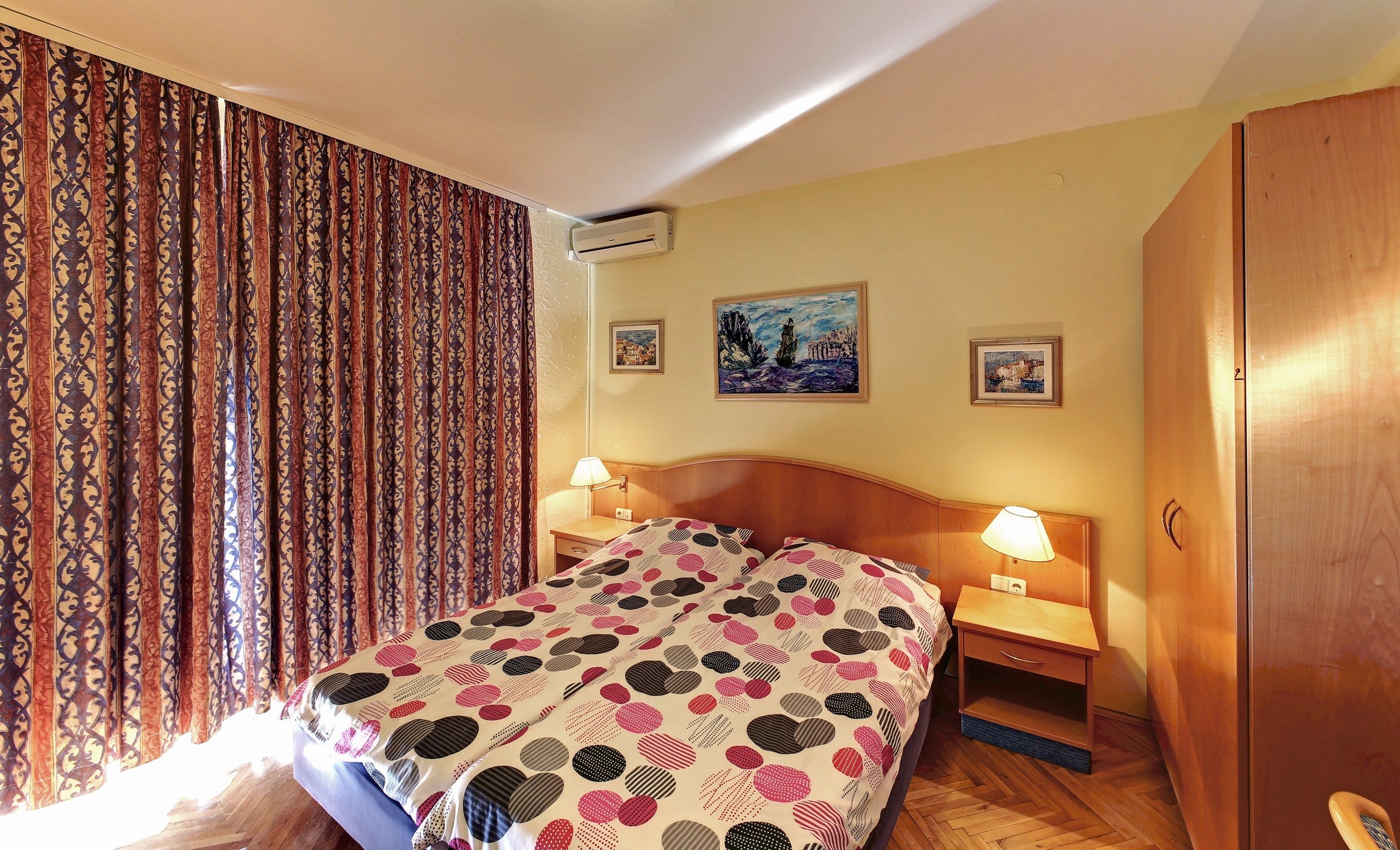 Une chambre d’hôtel comprenant un lit, deux tables de chevet avec des lampes, une armoire et deux tableaux encadrés accrochés au mur.
