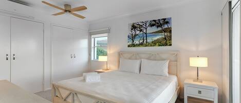 Quarto com cama, duas mesas de cabeceira, ventilador de teto, janela e um quadro na parede.