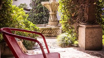 Un patio con una fuente de piedra, sillas de metal rojo y abundante vegetación.