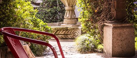 A courtyard with a stone fountain, red metal chairs, and lush greenery.