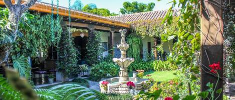 A garden with a fountain, lush greenery, and a building with a tiled roof.