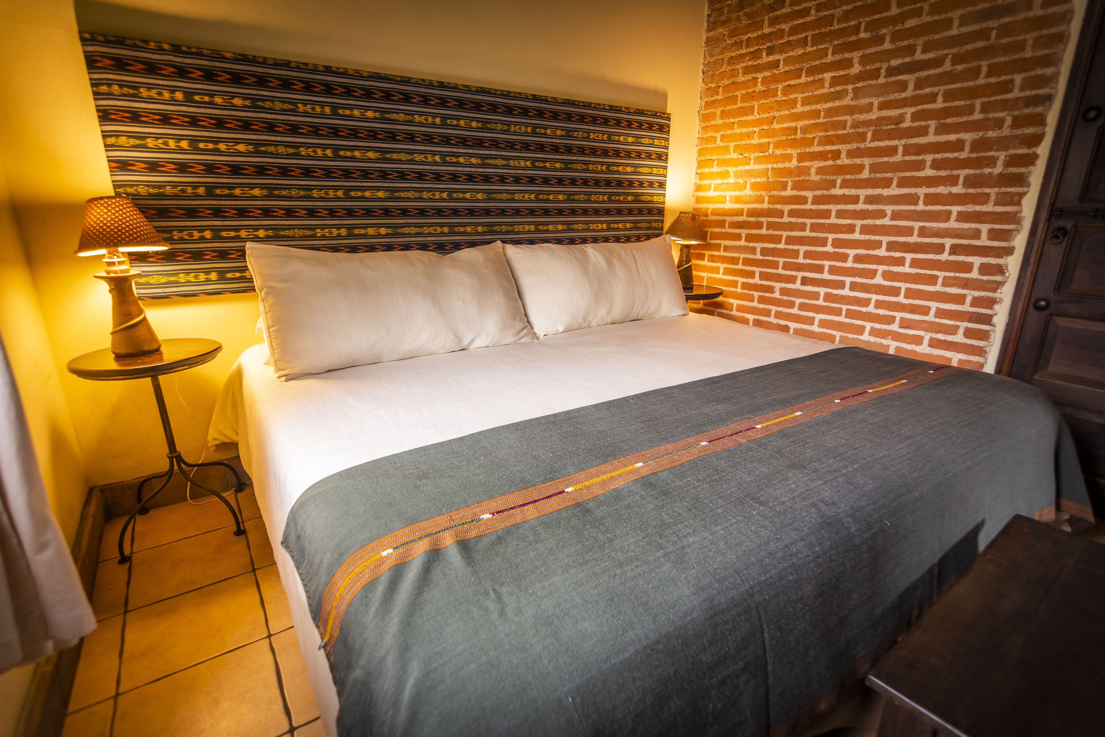 A neatly made bed with a grey bedspread and pillows, a patterned tapestry on the wall, and a brick wall to the right.