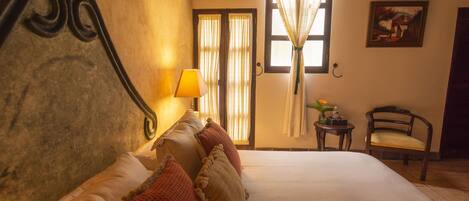 Una habitación de hotel con cama, una silla, una lámpara, ventana con cortinas y un cuadro en la pared.