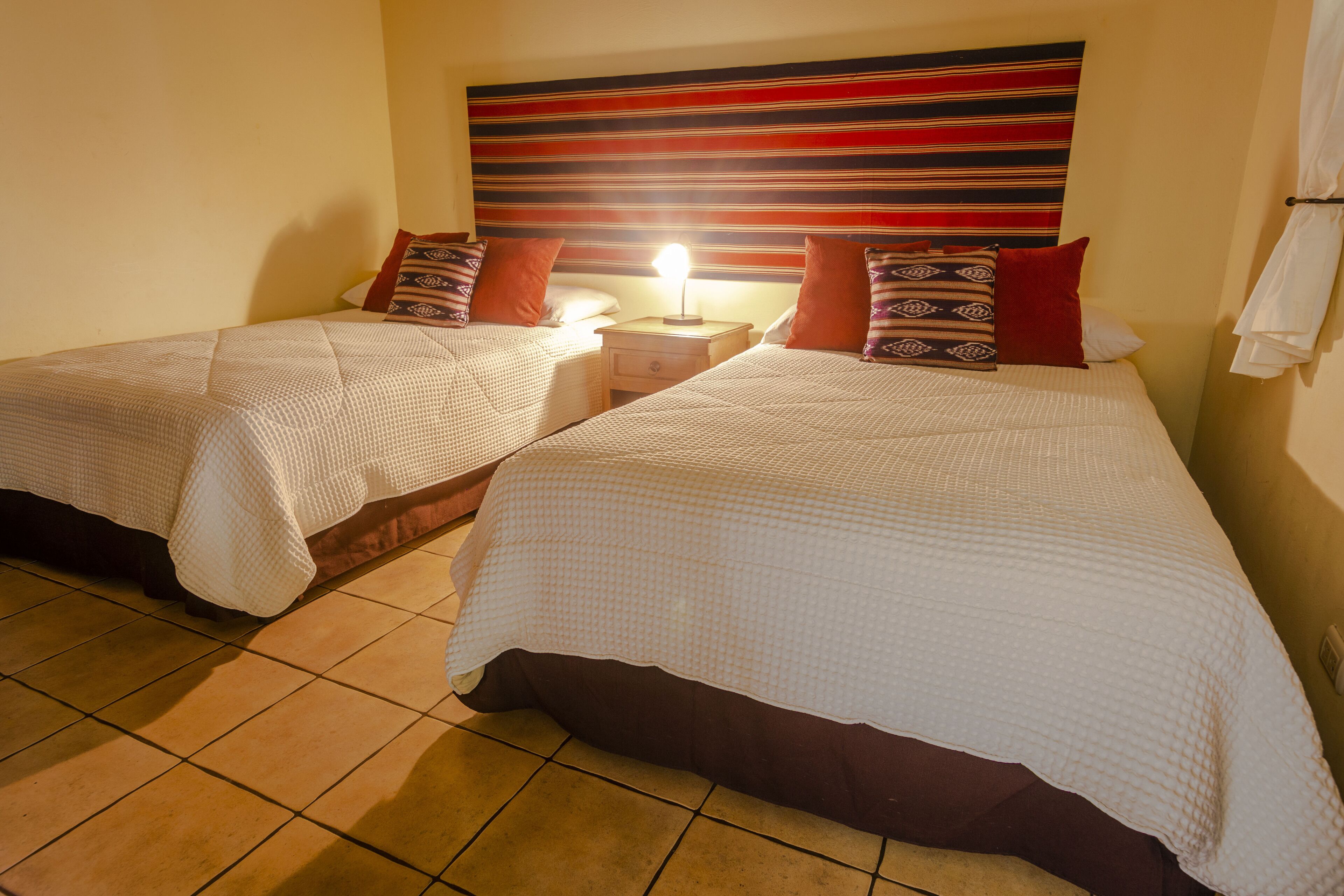 A hotel room with two beds, each with a white bedspread and a brown skirted bed base. There is a nightstand with a lamp between the beds, and a striped headboard above them.